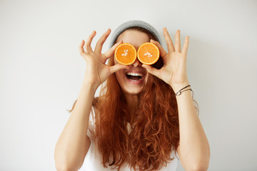Close up studio portrait of young smiling female holding halves of oranges at her eyes. Headshot of pretty girl with loose red hair in cap and white T-shirt having fun. Film effect, selective focus