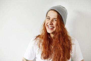 Portrait of cute redhead girl wearing gray winter cap and white T-shirt smiling with happiness and joy while posing against gray studio background. Headshot of cheerful female with happy expression