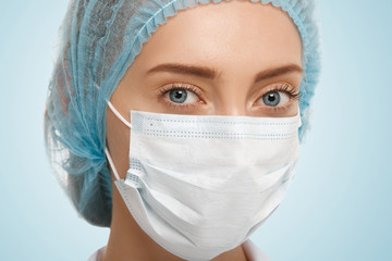 Close up view of cute Caucasian blue-eyed woman doctor wearing blue cap and white facemask looking at the camera isolated against studio background. Healthcare, medical and plastic surgery concept