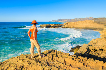 Young woman tourist standing on cliff and looking at beautiful sea bay, Fuerteventura island, Spain