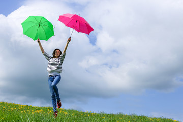 Une jeune fille saute de joie avec deux parapluies colorés dans un pré sur fond de ciel bleu et nuageux
