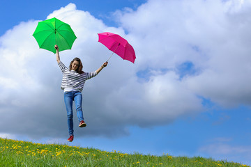 Une jeune femme semblant atterrir dans un pré grâce à deux parapluies sur fond de ciel bleu et nuageux