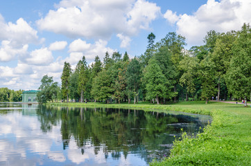 Fototapeta na wymiar Shore of a beautiful lake with a meadow and green forest backdrop and blue sky with clouds in the Gatchina Park in the vicinity of St. Petersburg.