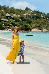 Beautiful blonde lady in long yellow tunic dress with her pretty little daughter in floral dress walking at seaside. They holding hands and smiling. Happy family on the beach. Happy mothers day.