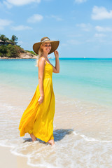 Carefree beautiful fashion blonde female in beach straw hat walking barefoot by sand of tropical shore and holding her long yellow dress. Natural woman beauty. Lady touch her hat and smiles.