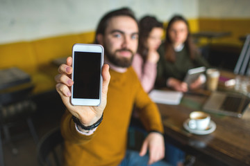 Young hiptser male showing smartphone on the camera in cafe