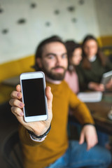 Young hiptser male showing smartphone on the camera in cafe