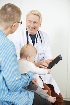 Male Pediatrician Showing Digital Tablet To Woman With Baby