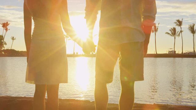 Romantic couple at sunset holding hands at beach watching the sun set at honeymoon vacation travel holidays. Woman man hand in hand at warm sunny evening with beautiful sunlight and flare.