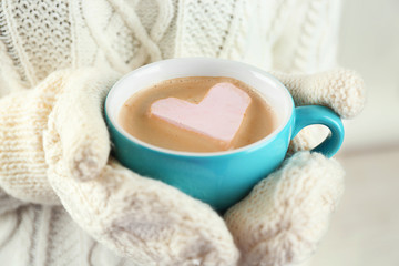 Female hands in warm mittens holding cup of hot cappuccino with heart marshmallow, close up