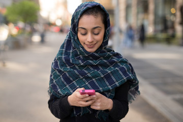 Young woman wearing hijab in city texting on cell phone