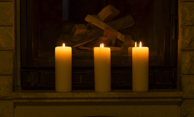 White large candles standing on the fireplace. Lighted candles on the stone fireplace with firewood. 
