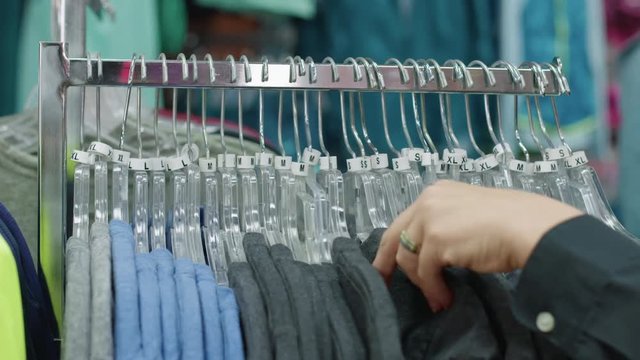 Woman's hands run from back to front across a rack of clothes on hangers, picking out a green T-shirt while browsing in a retail store.  Frontal view with rack focus.
