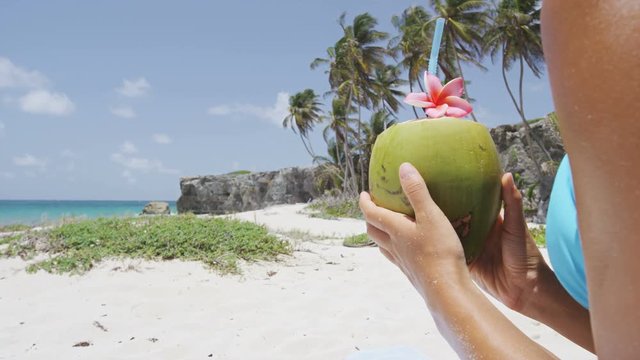 Fresh coconut water with straw on Caribbean beach in Barbados vacation. Closeup of unrecognizable woman holding young green tropical fruit for healthy snack during summer holidays.