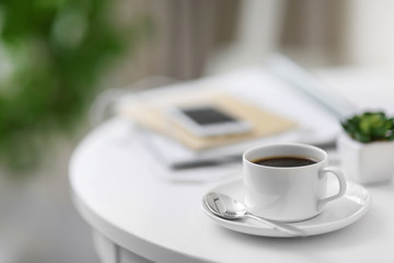 White cup of the coffee and a phone on the table in a light room.