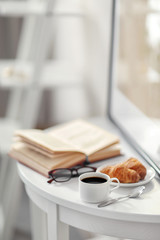 White cup of the coffee and an open book on the table  in a light room..