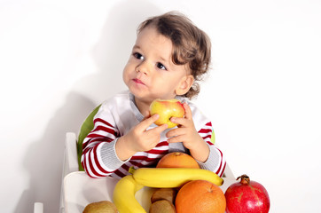 Fototapeta na wymiar Kid having a table full of organic food. Cheerful toddler eating healthy salad and fruits. Baby choosing between apples, bananas, oranges, avocado,pomegranate. kiwi. Isolated on white