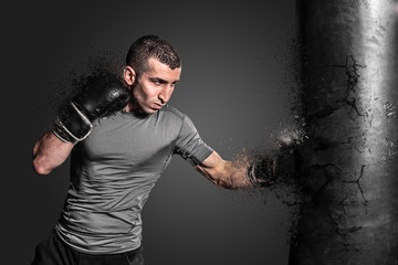 Boxer punching bag blow to the explosion. Young sportsman in black boxing gloves and gray uniforeme on a flat background.