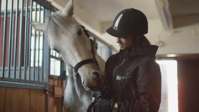 Young jockey girl is stroking a white horse in a stable. Shot on RED Cinema Camera.