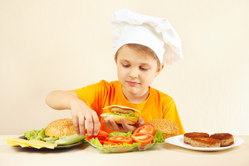 Little boy in chefs hat puts tomato on the hamburger