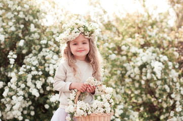 Fototapeta premium Smiling blonde kid girl holding basket with flowers outdoors. Looking at camera. Wearing floral wreath. 