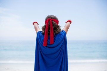 Rear view of boy in superhero costume with arms raised standing at sea shore