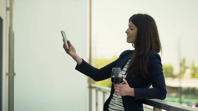 Happy woman with glass of wine taking selfie photo with cellphone on terrace
