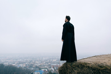 Priest bearded man raised his hands up on the hill above the town. Pastor posing outdoors in...
