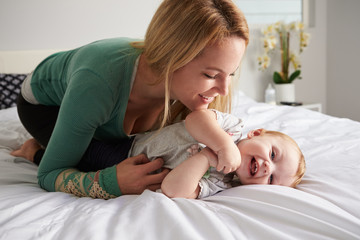 Obraz na płótnie Canvas Caucasian mother kneeling on bed, tickling her baby daughter