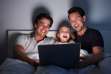 Young girl in bed watching laptop with her male gay parents