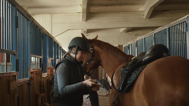 Young jockey girl is preparing a horse for a ride in stable. Shot on RED Cinema Camera.