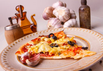 Homemade pizza out of the oven with ham, olives, tomato and cheese on baking paper. On the table in light wood. Pepper mill and spices, whole garlic.