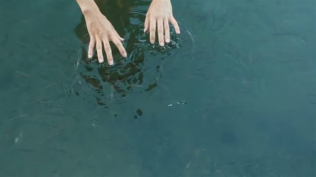 Reflection of beautiful naked girl in the mountain lake. Young woman's hands softly touch clean water surface with many little fishes in slow motion