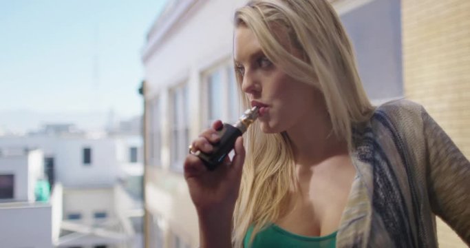 Close up of woman using an electronic cigarette on a balcony with background of city buildings.  Hand-held camera recorded at 60fps.