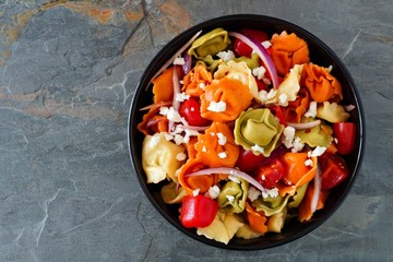 Colorful tortellini pasta salad with tomatoes and onions, overhead view on a dark slate background