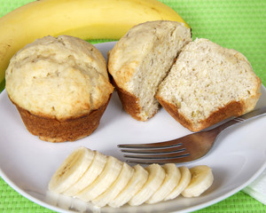 Banana bread muffins on a white plate with sliced bananas and fork on the plate, whole banana in background on green place mat, closeup