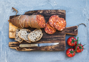 Wine snack set. Hungarian mangalica pork salami sausage, rustic bread and fresh tomatoes on dark wooden board over a rough grey-blue concrete background