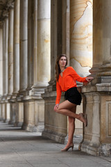 Beautiful blonde woman with orange blouse and short pants standing under classical arcardes