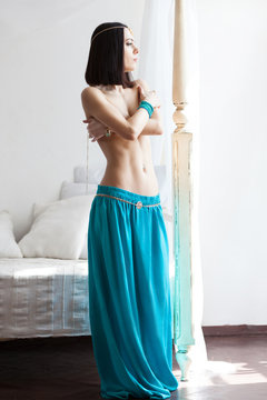 Attractive an slim woman in oriental garment and golden jewelry. She wears turquoise skirt and golden belt. She has a beautiful belly and perfect tanned skin. Bright sunny day, white room.
