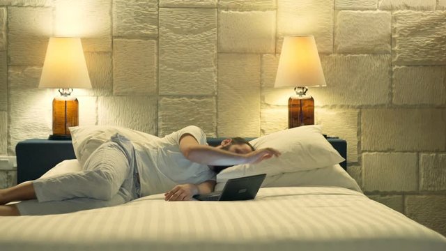 Young man using laptop and falling asleep on bed at night at home
