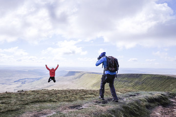 A photographer on a mountain taking a picture of a girl who is jumping on top , Pen Y Fan