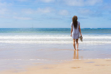 Fototapeta premium Young woman in dress and shoes in hand standing on beach and looking into sea.