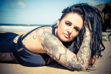 Heavily tattooed plus sized female model on South Shields beach.  She is wearing a swimsuit and has brown eyes.  She is lying on the sand.