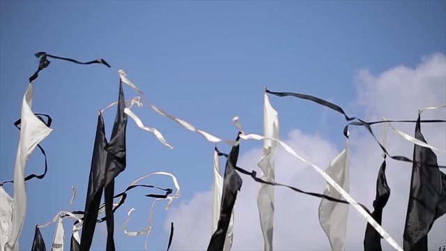 Black and white flags with long tapes sway on wind in summer day. Blue clear sky. Festival. Green trees. Slow motion. 