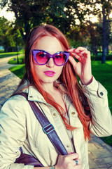 Portrait of Trendy Redhair Girl Standing in the park lane. Urban Fashion Concept  Red Lips and funny sunglasses.  Toned Color. Chic Long Hair. Colorized