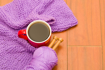 Closeup purple yarn ball with knitting needles and scarf in progress lying on desk, coffee mug sitting next to it, seen from above
