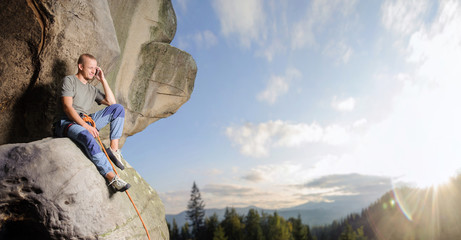 Athletic male climber is sitting on the big natural boulder secured with the rope against blue sky...
