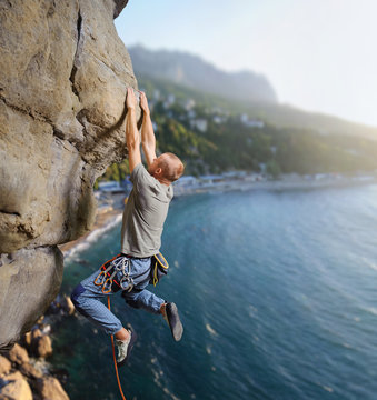 Side view of courageous man rock climber hanging on two hands and making difficult move up on overhanging cliff against scenic sea coast background. Climbing equipment. Summer time