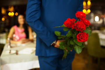 young guy holds behind flowers, red roses, meeting a young couple in a restaurant. Focus on flowers