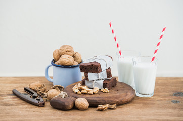 Chocolate brownie slices wrapped in paper and tired with rope, milk glasses, stripe straws, enamel mug of walnuts on rustic wooden table, white background